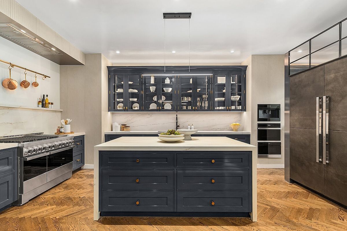 This is a well-lit, modern kitchen featuring dark blue cabinetry with glass-fronted upper cabinets displaying dishware. A large island with a white countertop and dark blue base sits in the center, complemented by stainless steel appliances and a herringbone wood floor. The kitchen exudes a sophisticated and functional design.