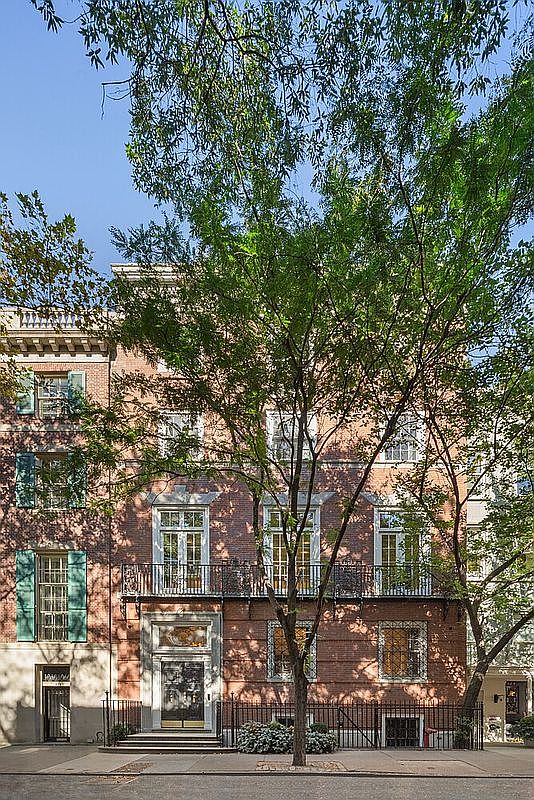 This is a front view of a stately brick townhouse. The building features multiple stories with large windows, some with shutters, and wrought iron balconies. A tree partially obscures the facade, adding a touch of nature to the urban setting.