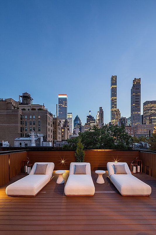 This image showcases a luxurious rooftop deck with stunning city views. Three modern lounge chairs with small side tables are arranged on a wooden deck, creating an inviting space for relaxation. The backdrop features a cityscape at dusk, adding to the upscale and serene ambiance.