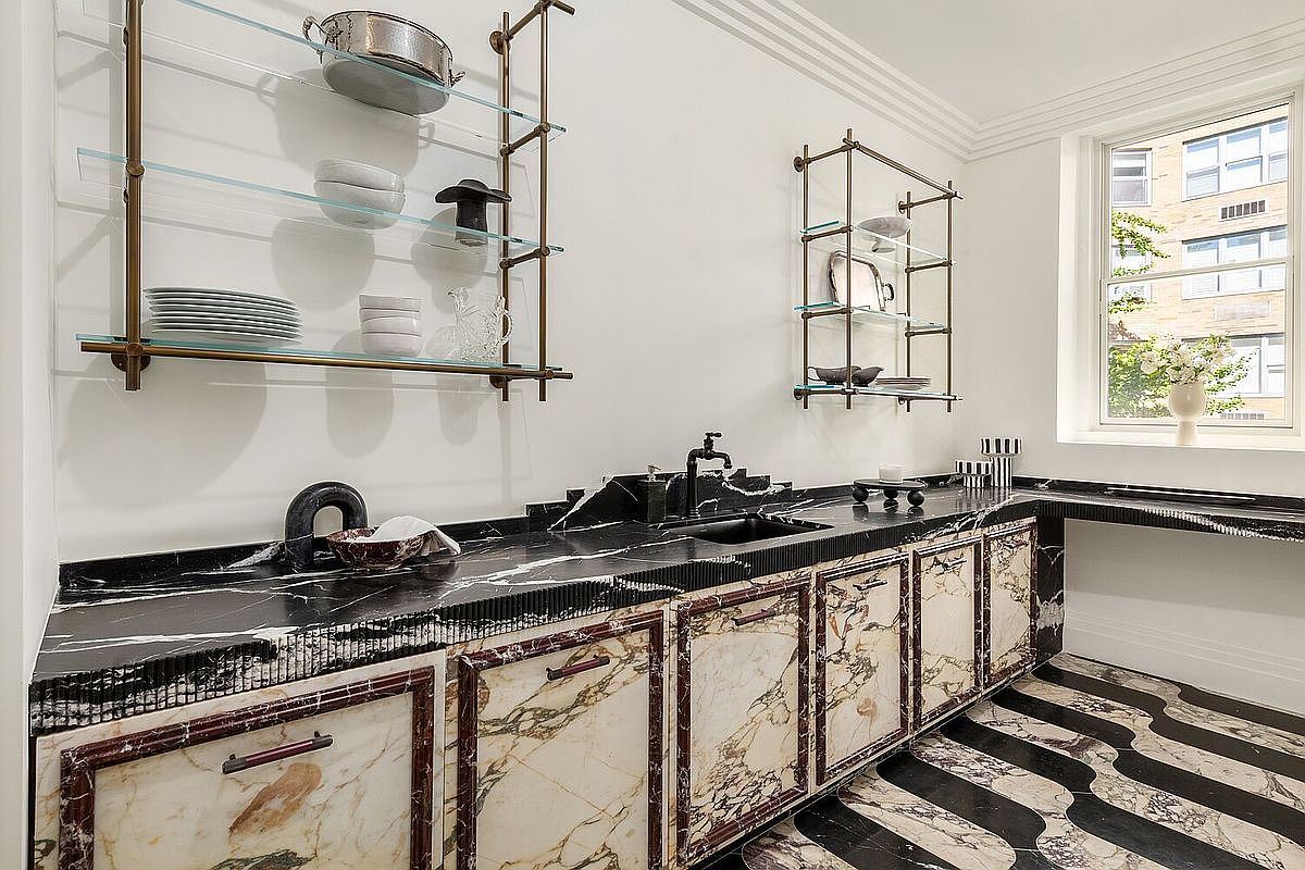 This interior shot showcases a unique kitchen design featuring marble countertops and cabinet fronts with dark wood trim. Open shelving displays dishware, while a window provides natural light. The black and white patterned floor adds a bold, artistic touch to the space.