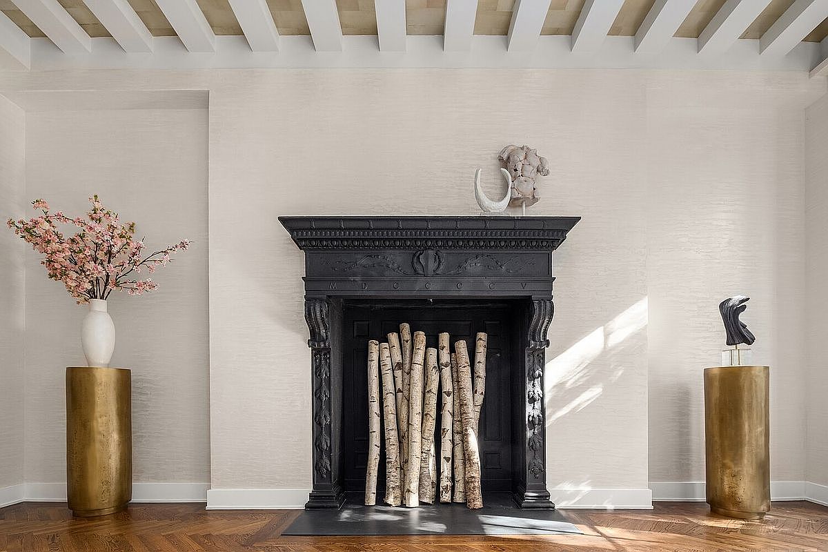 The image showcases a sophisticated living room interior featuring a black ornate fireplace as a focal point, adorned with birch logs. Flanking the fireplace are two golden cylindrical pedestals, one supporting a vase with pink blossoms and the other a modern sculpture. The room is bathed in natural light, highlighting the herringbone wood flooring and the textured off-white walls, creating an elegant and inviting atmosphere.