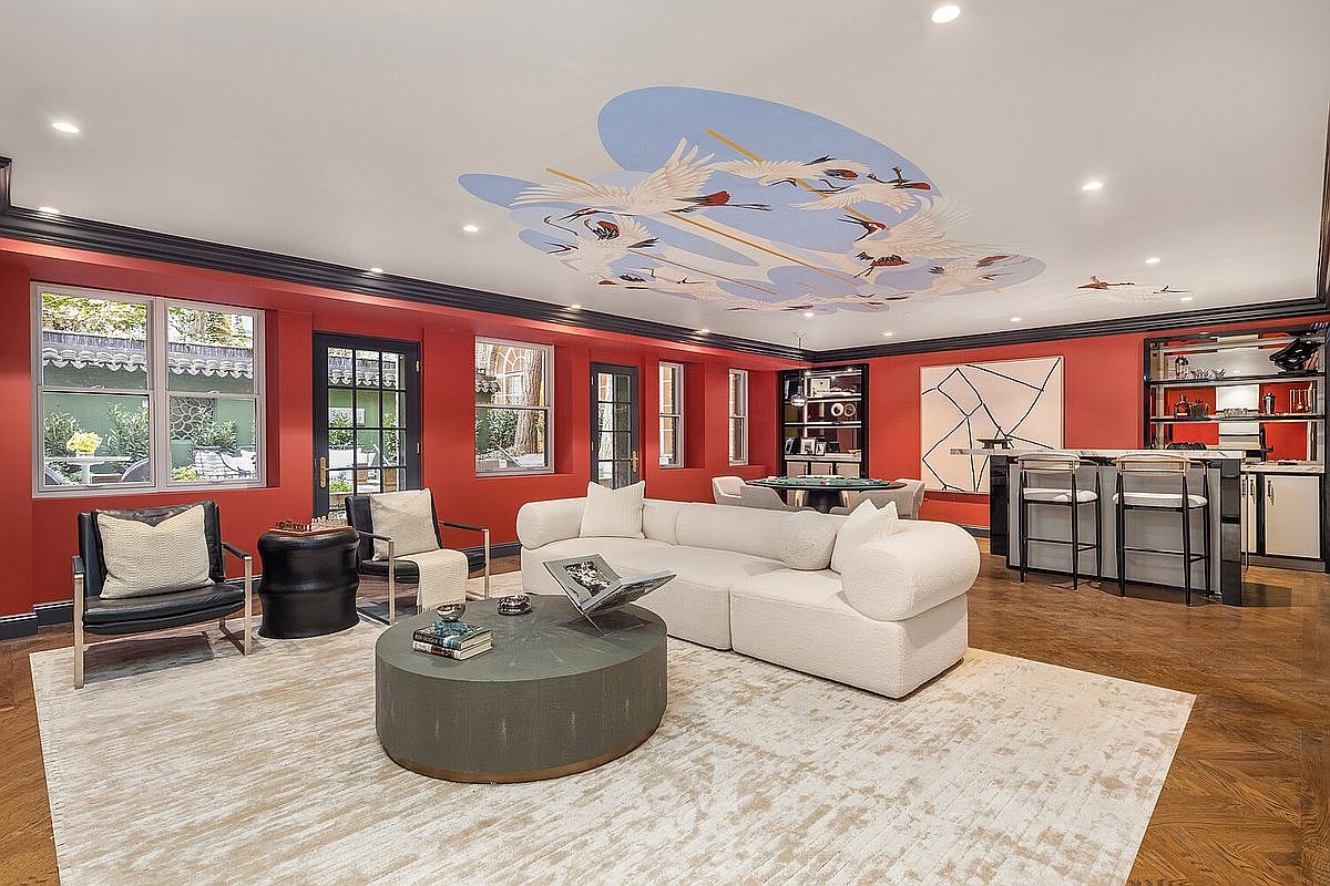 This is a stylish living room featuring red walls with dark trim, a unique ceiling mural, and hardwood floors covered by a large area rug. The room is furnished with a modern white sectional sofa, a round coffee table, and two armchairs, creating a comfortable and inviting space. A bar area with stools is visible in the background, adding to the room's functionality and entertainment potential.