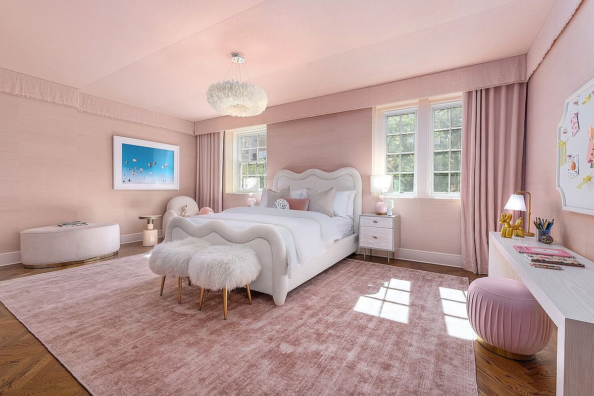 This is a charming bedroom featuring a unique white bed with a wavy headboard and footboard, complemented by fluffy white stools. The room is bathed in a soft pink hue, from the walls and ceiling to the plush rug, creating a warm and inviting atmosphere. Natural light streams in through the windows, highlighting the room's elegant and whimsical design.