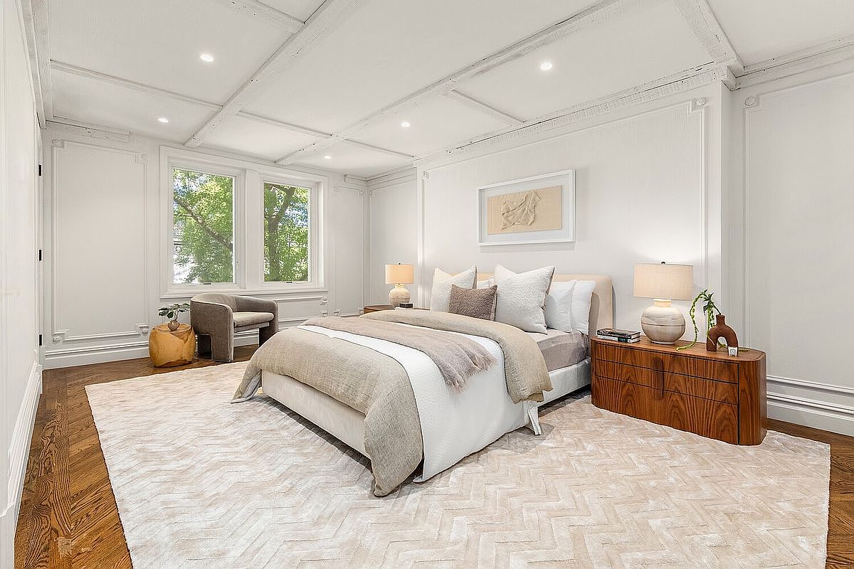 This is a bright and airy primary bedroom featuring a large bed with neutral-toned bedding, flanked by wooden nightstands and lamps. The room is decorated in a classic style with white walls, detailed molding, and a coffered ceiling. A comfortable chair sits near the windows, which provide natural light and a view of the outdoors.