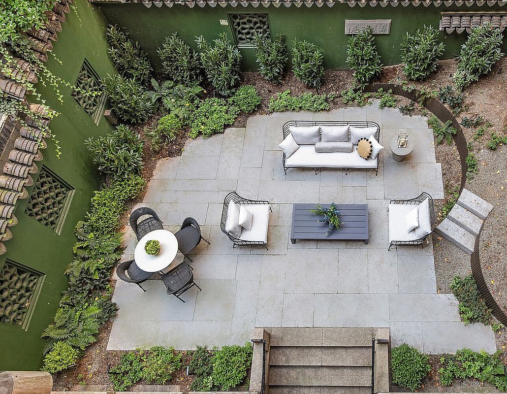 This aerial view showcases a meticulously designed courtyard garden. The space features a stone-paved patio with outdoor seating, including a sofa, chairs, and a coffee table. Lush greenery surrounds the patio, creating a serene and private outdoor living area, perfect for relaxation and entertaining.