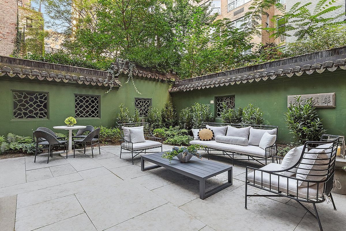 This image showcases a beautifully designed outdoor patio area, featuring stylish patio furniture including a sofa, chairs, and a coffee table, all arranged on a stone-paved surface. The patio is enclosed by a unique green wall with decorative windows and a tiled roof, creating a private and serene outdoor living space. Lush greenery surrounds the patio, enhancing the tranquil atmosphere and adding to the overall appeal of the property.