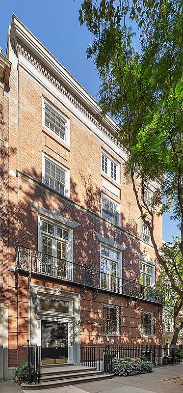 This is a stately, multi-story brick townhouse with a classic architectural style. The facade features symmetrically arranged windows, a wrought-iron balcony, and an elegant entryway with a prominent door. Mature trees partially frame the building, adding to its curb appeal and suggesting a well-established neighborhood.