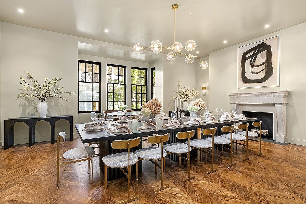 This is a well-lit dining room featuring a long, dark-colored dining table set for a formal meal, surrounded by modern chairs with gold frames and light-colored cushions. A striking chandelier hangs above the table, and a large abstract painting adorns the wall above a fireplace. The room also includes a dark console table and large windows, all contributing to an elegant and sophisticated atmosphere.