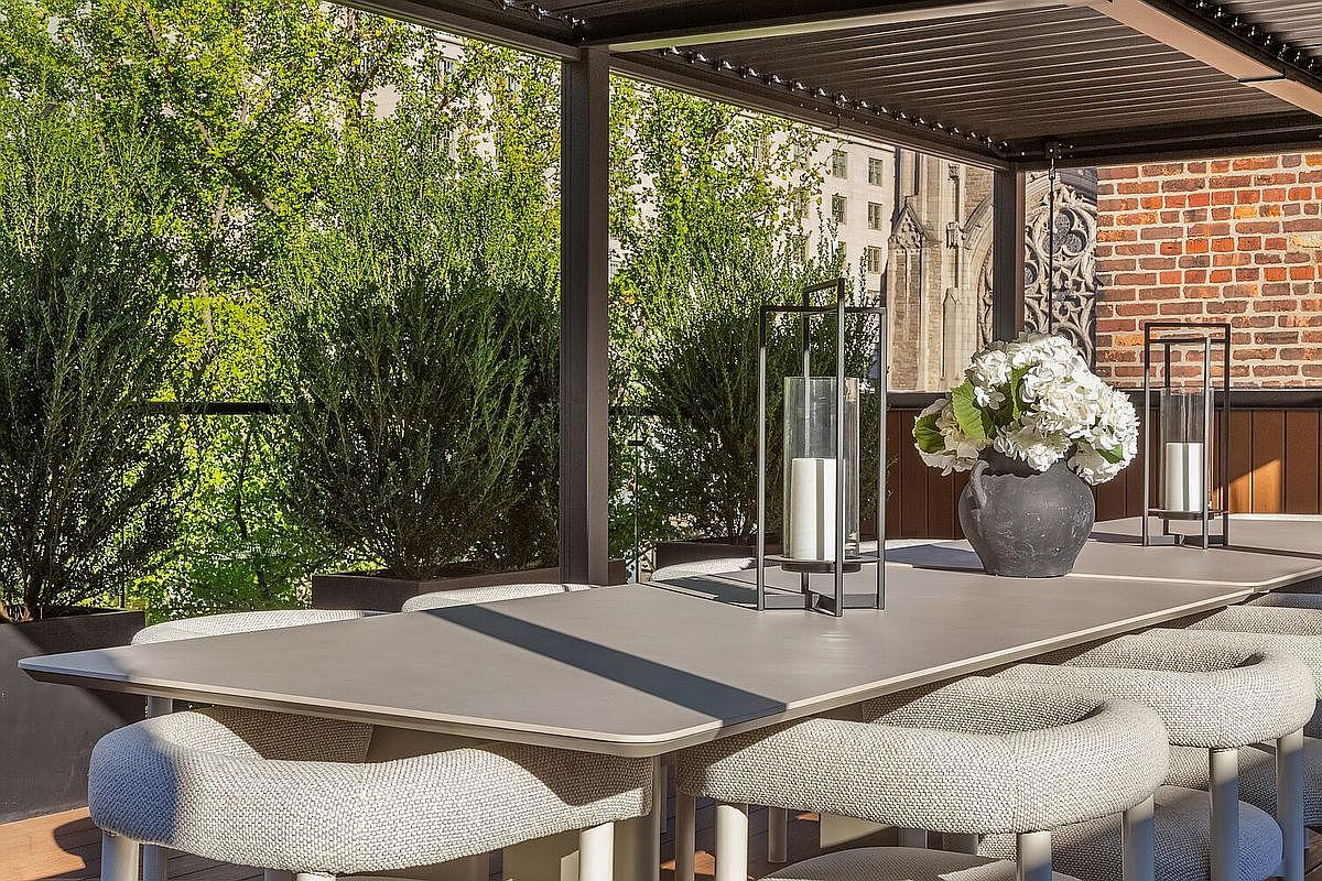 This image showcases a stylish outdoor patio or balcony area, featuring a large dining table with modern chairs, decorative lanterns, and a floral centerpiece. The space is partially covered by a pergola, offering shade, while lush greenery in the background adds a sense of privacy and tranquility. The view includes a glimpse of a building, suggesting an urban setting.