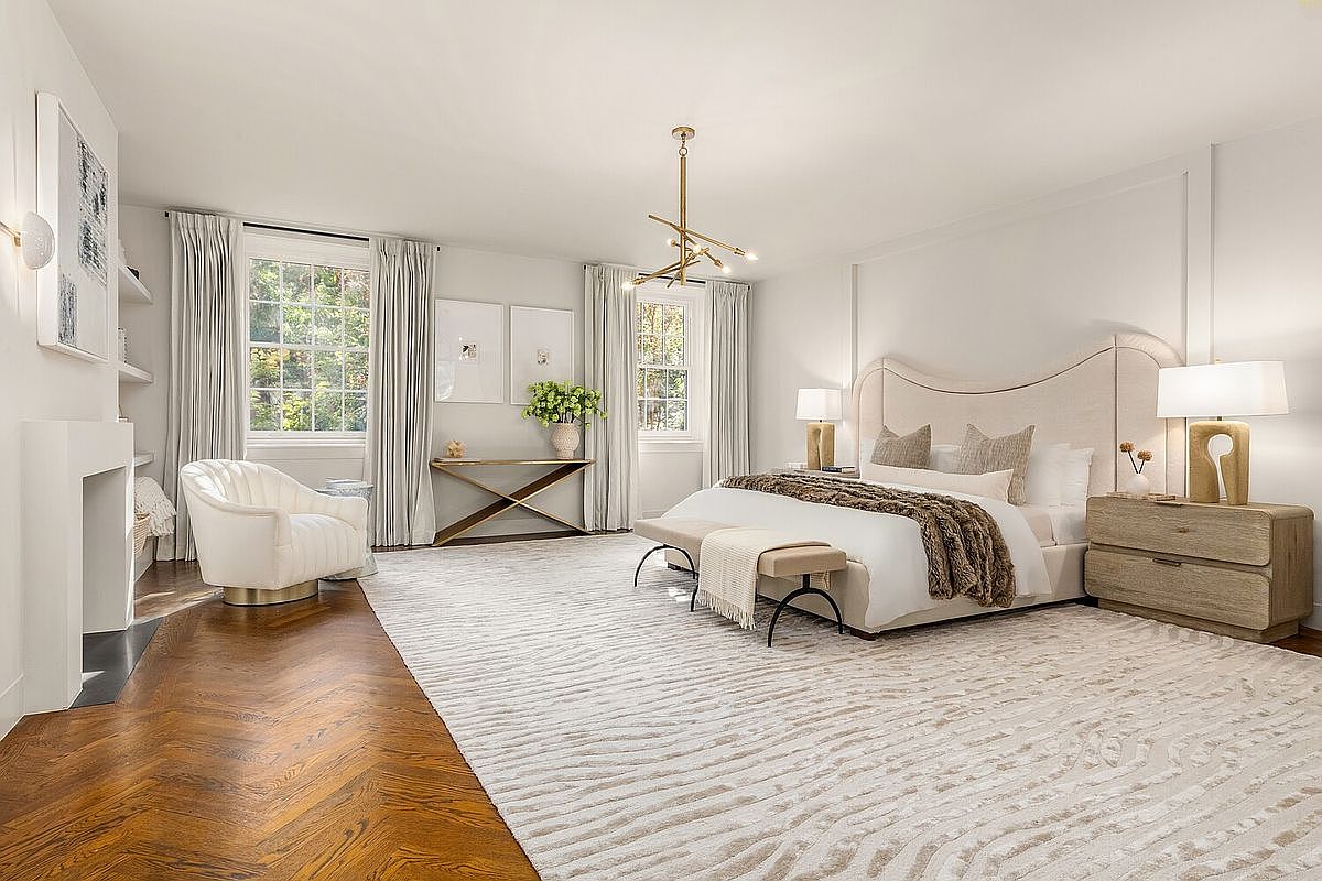 This is a spacious primary bedroom featuring a large bed with a curved headboard, a neutral-toned rug with a subtle pattern, and hardwood floors. The room is well-lit with natural light from the windows and complemented by stylish bedside lamps. The overall impression is one of luxury and comfort.