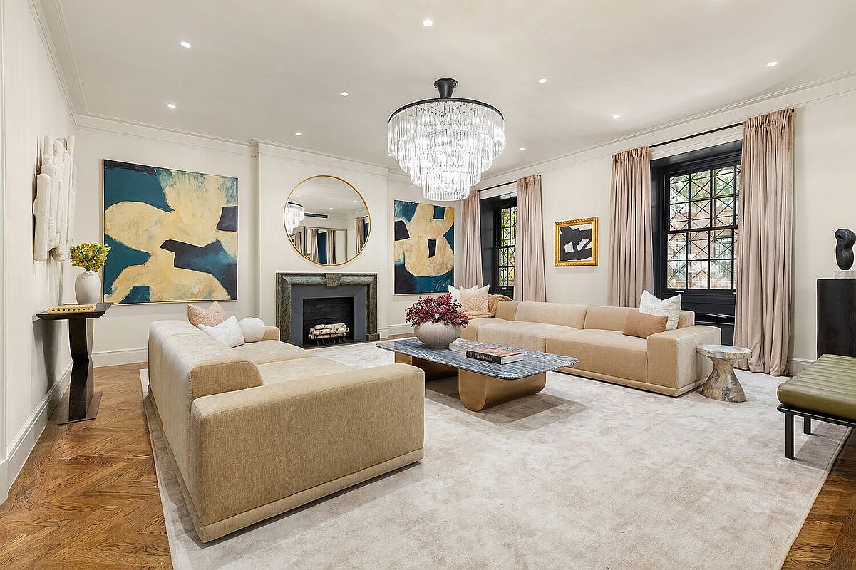 This is a well-lit living room featuring two beige sofas, a marble coffee table, and a large chandelier. The room is decorated with abstract art and a round mirror above a fireplace, creating a sophisticated and inviting atmosphere. Large windows with elegant drapes add to the room's charm and provide natural light.