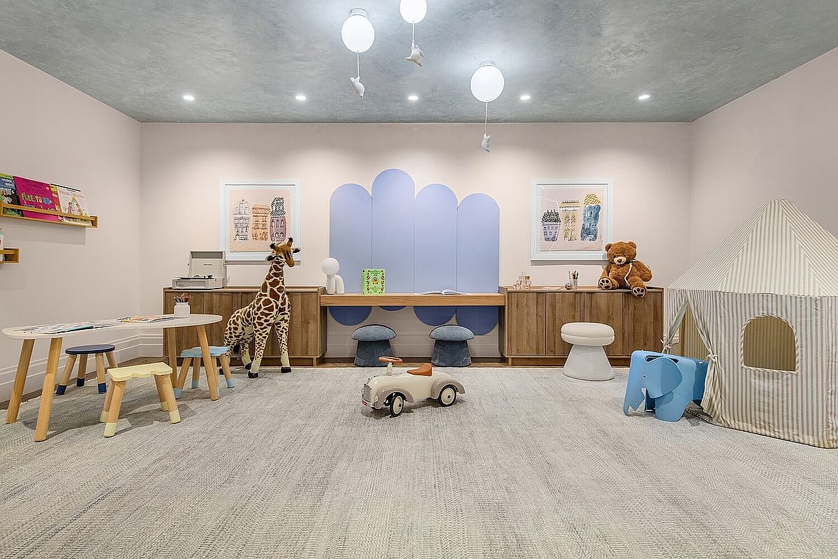 This is a charming children's playroom featuring a light gray carpet, light pink walls, and a variety of playful elements. The room includes a round table with small stools, a giraffe plush, a toy car, and a striped tent, creating a whimsical and inviting space for kids to play and explore. The perspective is a wide shot, showcasing the entire room and its layout.