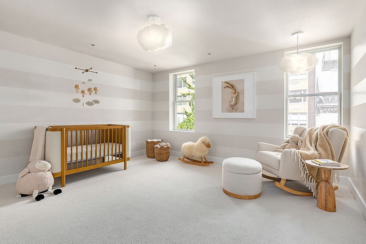This is a serene nursery featuring a wooden crib with a soft blanket, a rocking chair with a throw, and a whimsical rocking sheep. The room is decorated with horizontal stripes in neutral tones, creating a calming atmosphere. Natural light streams in through the windows, enhancing the room's peaceful ambiance.