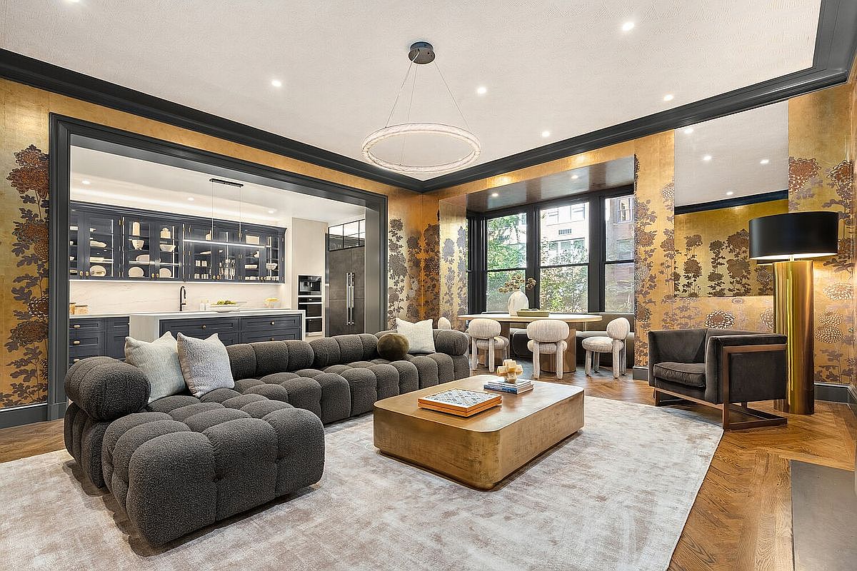 This is a luxurious living room featuring a large, modular gray sofa, a gold-toned coffee table, and a patterned rug. The walls are adorned with decorative wallpaper, and there's an open view into a modern kitchen with dark cabinetry. A dining area is visible by the window, creating an open and inviting space.
