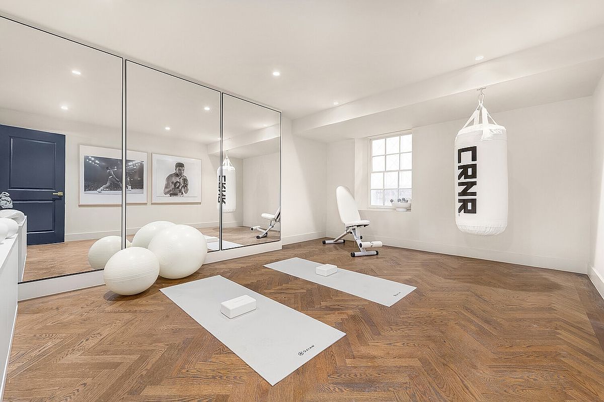This is a well-appointed home gym featuring herringbone wood floors, mirrored walls, and ample natural light from a window. The space includes exercise balls, yoga mats, a weight bench, and a punching bag, creating a functional and stylish workout area. The perspective is from the entrance of the room, showcasing the layout and equipment.