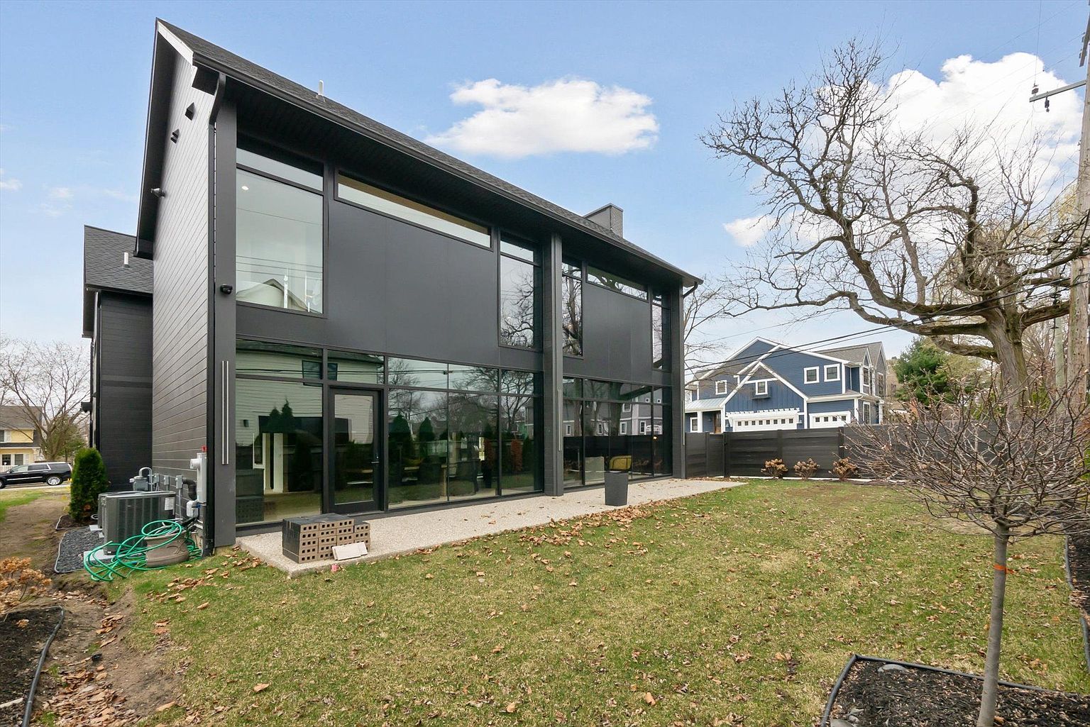This is a rear view of a modern, two-story house with a striking black exterior and large windows. The backyard features a well-maintained lawn with scattered leaves, a small tree, and a dark wooden fence. A blue house is visible in the background, adding depth to the scene.