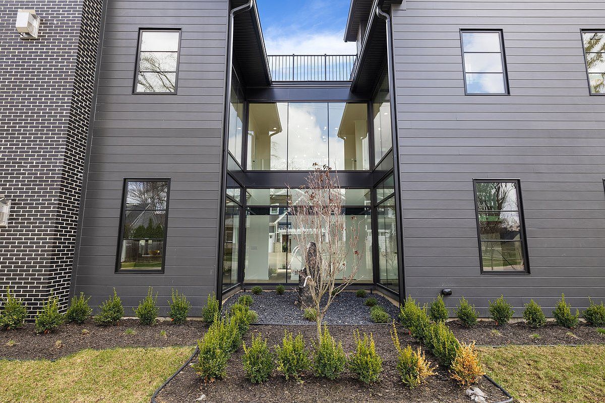 This is a striking front view of a modern home featuring a unique glass-enclosed entryway connecting two wings of the house. The exterior is clad in gray siding, complemented by black window frames and a dark brick accent wall. The landscaping includes neatly arranged shrubs and a small tree in a bed of dark gravel, adding to the property's curb appeal.