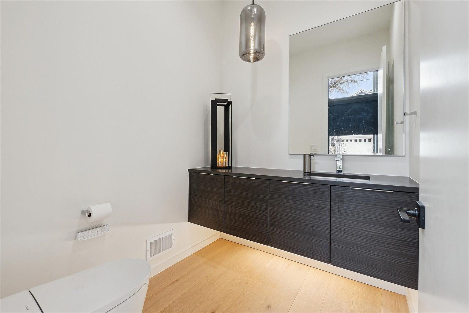 This is a modern guest bathroom featuring a floating dark wood vanity with a gray countertop and a rectangular mirror above. A sleek pendant light hangs above the vanity, and a toilet with a paper holder is visible on the left. The light wood flooring adds warmth to the space, creating a clean and contemporary aesthetic.