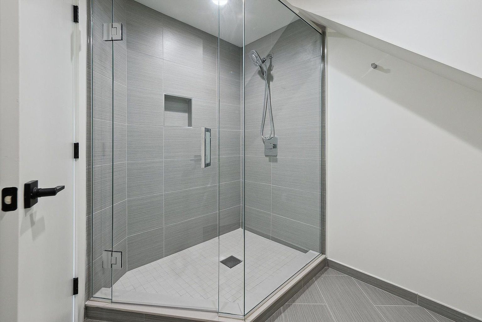 This is a modern bathroom featuring a glass-enclosed shower with gray, horizontally-laid tiles. The shower includes a built-in niche and a handheld shower head. The floor outside the shower has matching gray tiles, while the shower floor has smaller, square tiles. A black door handle is visible on the left.