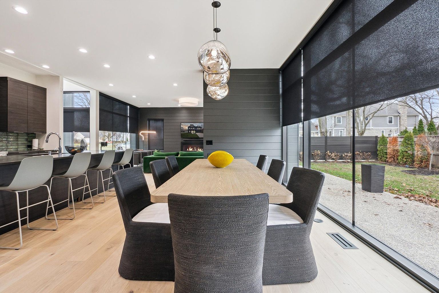 This is a modern dining room featuring a long wooden table surrounded by gray upholstered chairs. Above the table hangs a unique, multi-globe pendant light fixture. Large windows offer a view of the backyard, and the room seamlessly connects to the kitchen and living areas, creating an open and inviting space.