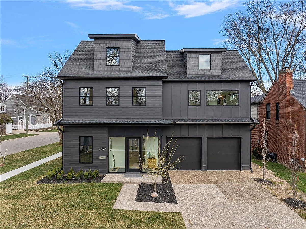 This is a front exterior view of a modern, two-story house with a dark gray facade. The house features a symmetrical design with two dormers on the roof, a two-car garage, and a well-manicured lawn. The entryway is accented by a small tree and minimalist landscaping, creating a sophisticated and inviting curb appeal.