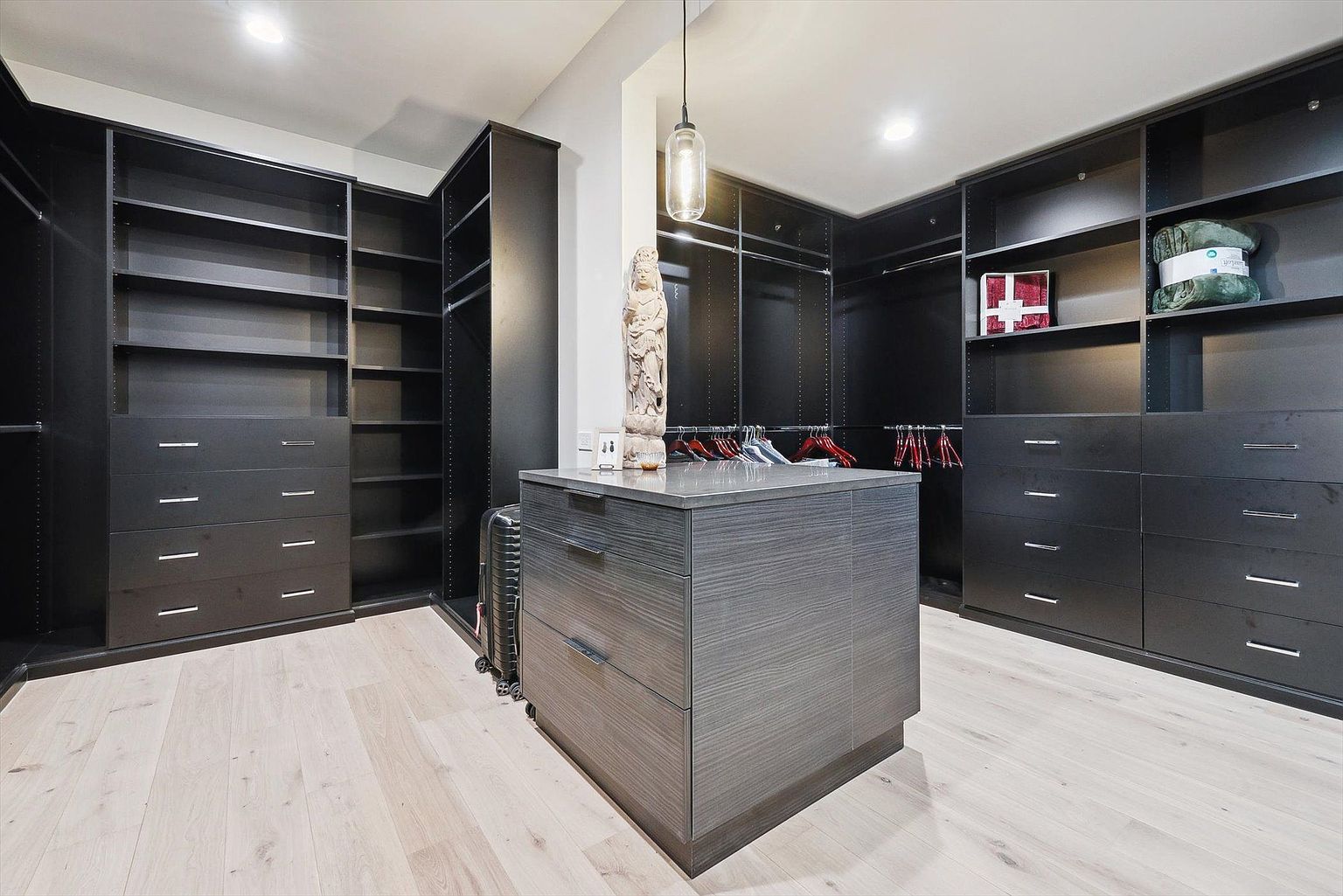 This is a well-organized walk-in closet featuring dark-colored shelving and drawers, providing ample storage space. A central island with a gray wood-grain finish offers additional surface area and drawer space. The closet is illuminated by recessed lighting and a pendant light, creating a sophisticated and functional space.