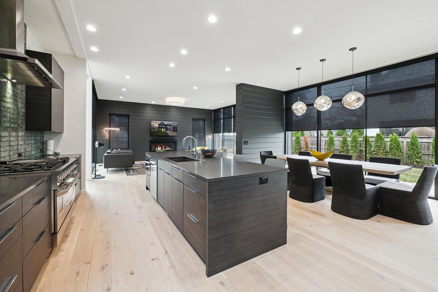 This is a modern, open-concept kitchen and dining area featuring dark wood cabinetry and a large central island with a sink. The kitchen flows into a dining space with a long table and modern pendant lighting, with large windows offering natural light and views of the exterior. The overall impression is sleek, contemporary, and spacious, ideal for entertaining.