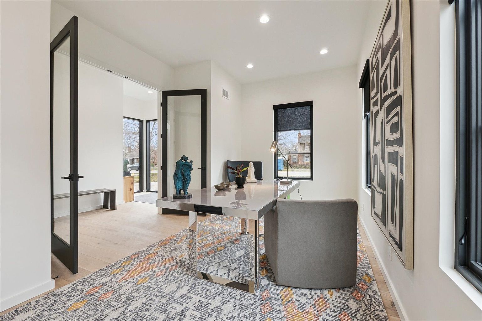 This is a modern home office featuring a sleek, reflective desk with a gray chair. The room has white walls, black-framed windows, and a stylish rug with geometric patterns. A sculpture sits on the desk, adding an artistic touch to the space.