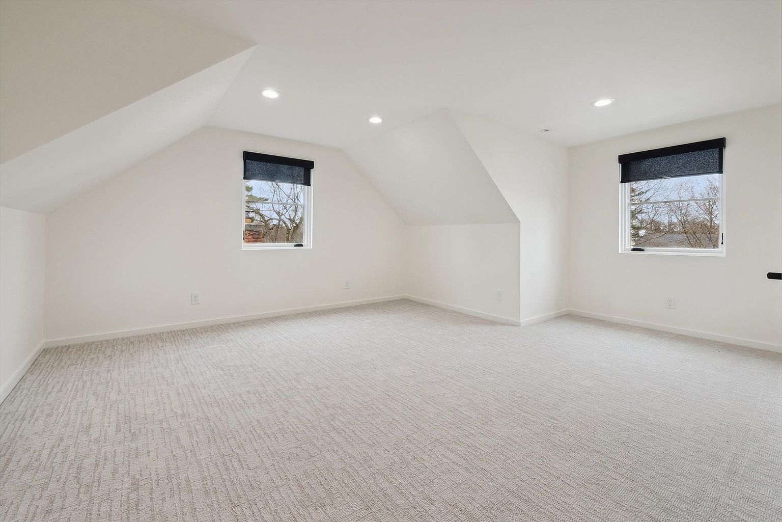 This is a bright and spacious bedroom featuring a neutral color palette with white walls and light gray carpeting. The room has two windows with dark blinds, allowing for ample natural light. The sloped ceilings add architectural interest, making it a cozy yet open space, ideal for a guest room or additional living area.