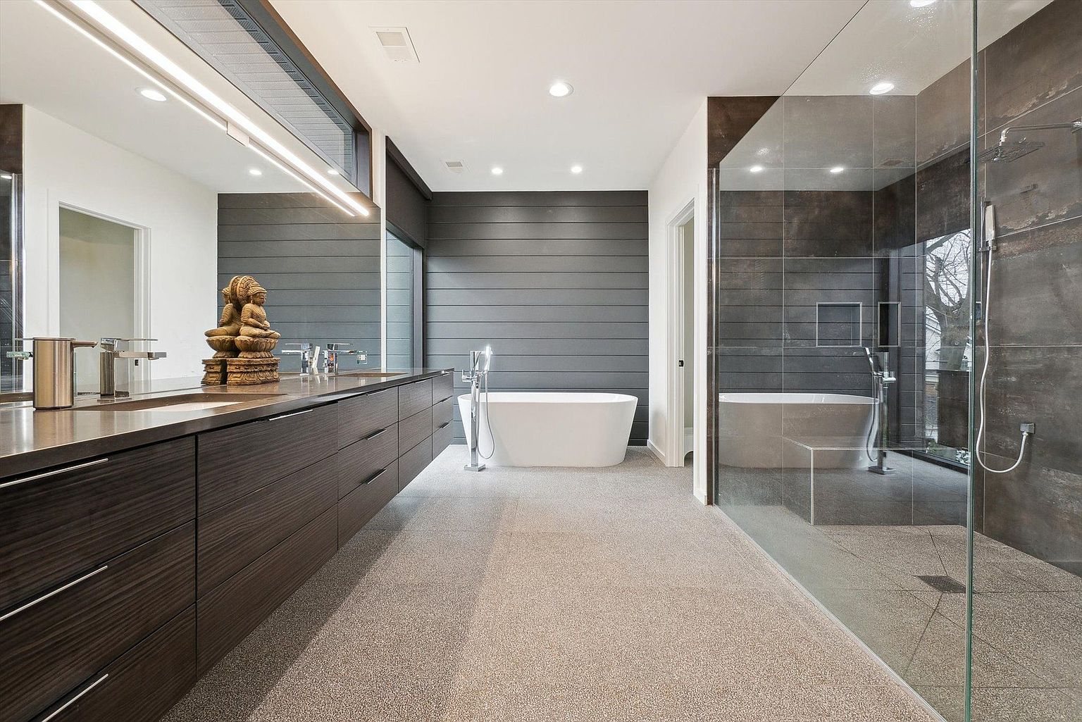 This is a modern primary bathroom featuring a long, dark wood vanity with dual sinks and a decorative statue. A freestanding white bathtub sits against a gray horizontal plank wall, and a glass-enclosed shower with dark tile is visible on the right. The flooring is a light speckled material, and the overall design is sleek and contemporary.