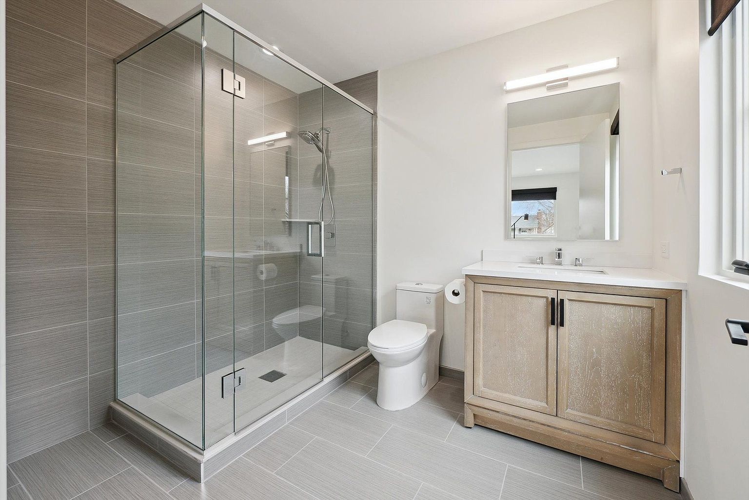 This is a well-lit primary bathroom featuring a glass-enclosed shower with gray tiled walls, a white toilet, and a vanity with a light wood cabinet and white countertop. A rectangular mirror hangs above the vanity, reflecting natural light. The floor is covered in gray tiles, creating a cohesive and modern aesthetic.
