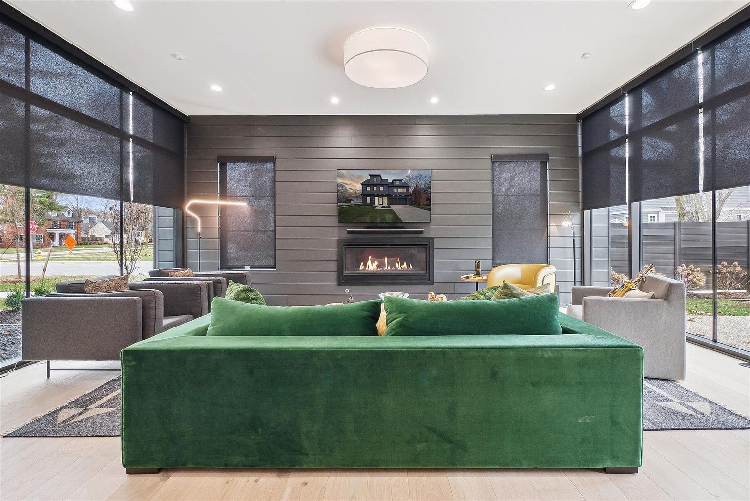 This is an interior shot of a modern living room featuring a large green velvet sofa as the focal point. The room includes a fireplace with a television mounted above it, flanked by windows with dark shades. The overall aesthetic is contemporary and stylish, with a focus on comfort and design.