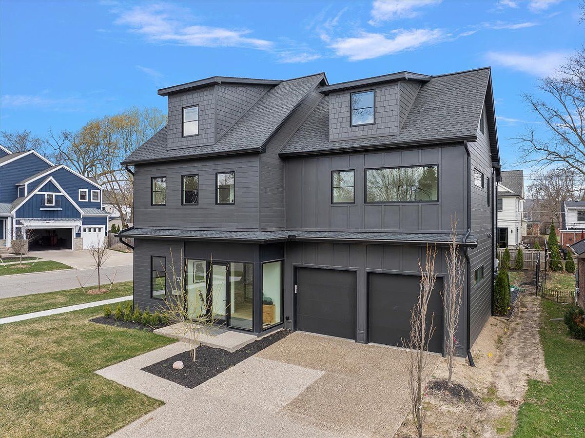 This is an aerial view of a modern two-story house with a dark gray exterior and a matching roof. The house features a two-car garage, a small front yard with landscaping, and a contemporary design with clean lines and large windows. The perspective highlights the architectural details and the overall curb appeal of the property.