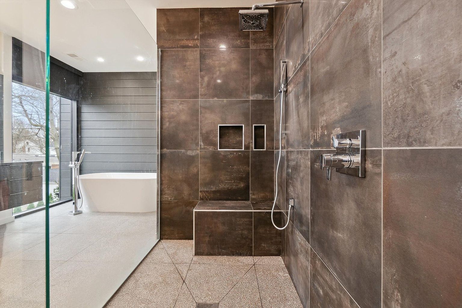 This is a modern primary bathroom featuring a walk-in shower with dark, large-format tiles and a built-in bench. The shower has a rainfall showerhead and a handheld sprayer. Through a glass partition, a freestanding bathtub is visible, set against a backdrop of horizontal wood paneling and a large window, creating a spa-like atmosphere.