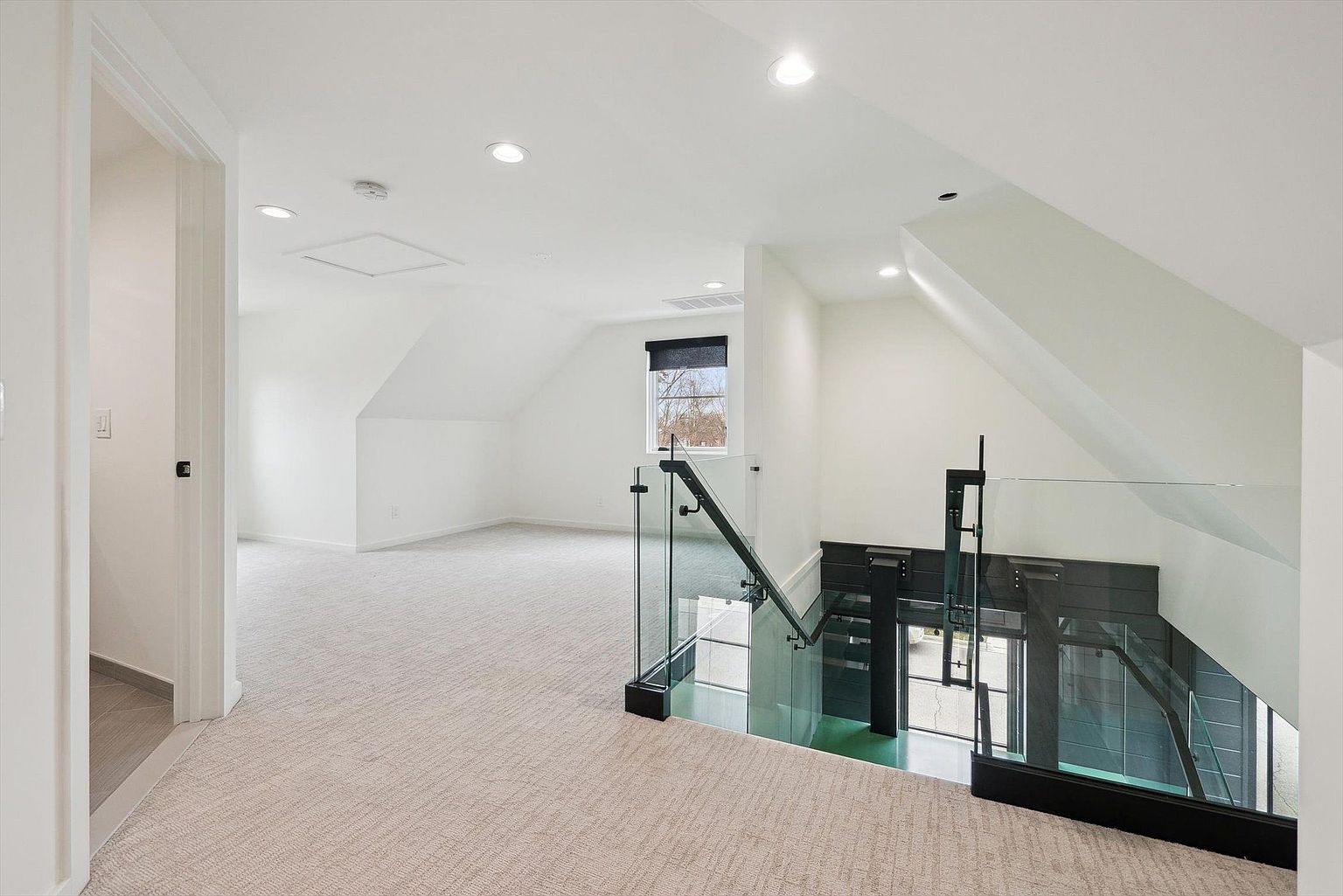 This interior shot showcases a bright, modern hallway with a staircase featuring glass railings and black accents. The space is well-lit with recessed lighting and natural light from a window, highlighting the clean lines and minimalist design. The light-colored carpet adds warmth and texture to the area.