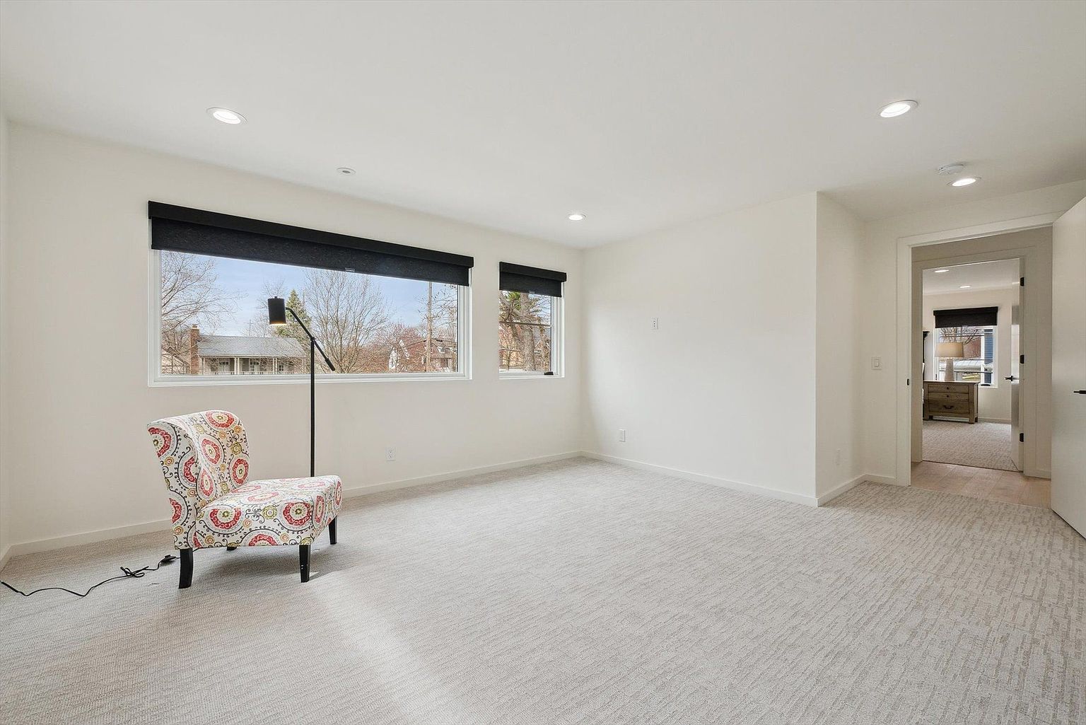 The image showcases a bright and airy bedroom with neutral-toned carpet and white walls, creating a spacious feel. Natural light streams in through two windows fitted with dark blinds, complemented by recessed lighting in the ceiling. A patterned accent chair and a modern floor lamp add a touch of style, while an open doorway reveals a glimpse into an adjacent room, suggesting a well-connected layout.