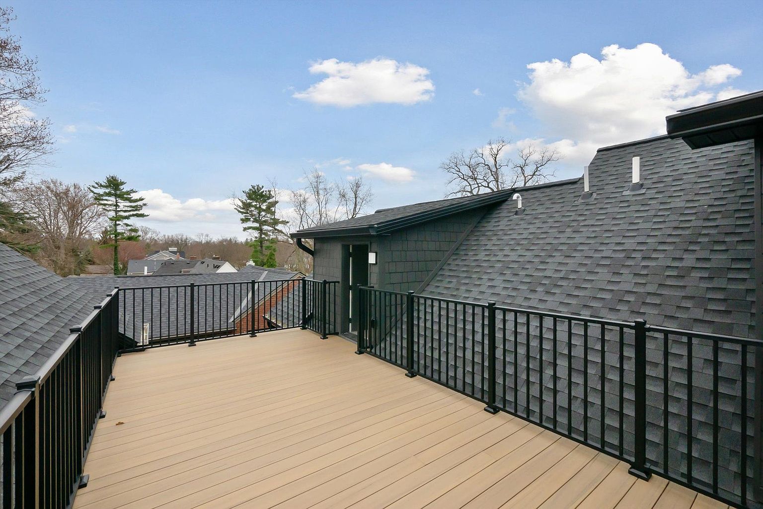 This image showcases a spacious outdoor deck or balcony area, featuring a light-colored wooden floor and a black metal railing. The deck is adjacent to a building with dark gray siding and a dark shingled roof. The view extends to other rooftops and trees, with a partly cloudy sky above, creating an inviting outdoor living space.