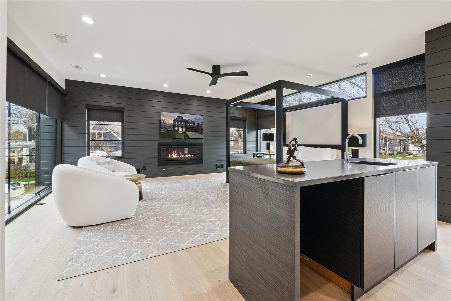 This is a spacious primary bedroom featuring a modern design with dark wood paneling and light wood flooring. The room includes a fireplace with a television mounted above, a seating area with white armchairs, and a four-poster bed. A kitchen island with a sink and modern fixtures is also present, creating a unique and luxurious living space.