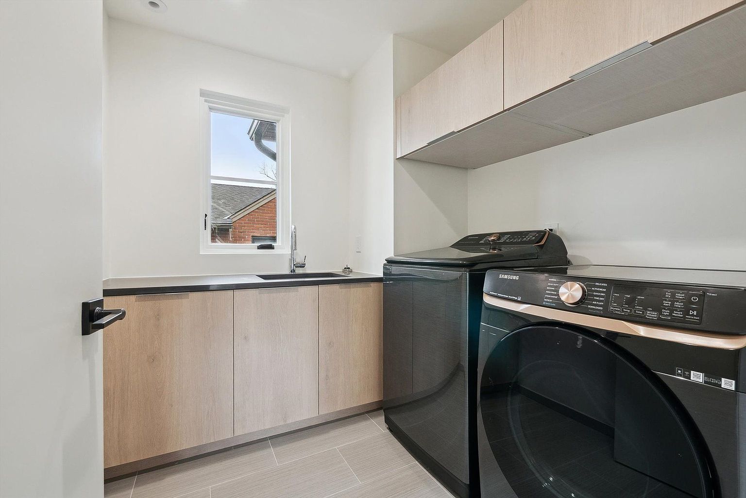 This is a well-lit laundry room featuring modern, light wood cabinetry with a dark countertop and a stainless steel sink beneath a window. A dark-colored Samsung washer and dryer set are positioned against the wall, complemented by additional light wood cabinets above. The room has a clean and functional design, perfect for a modern home.