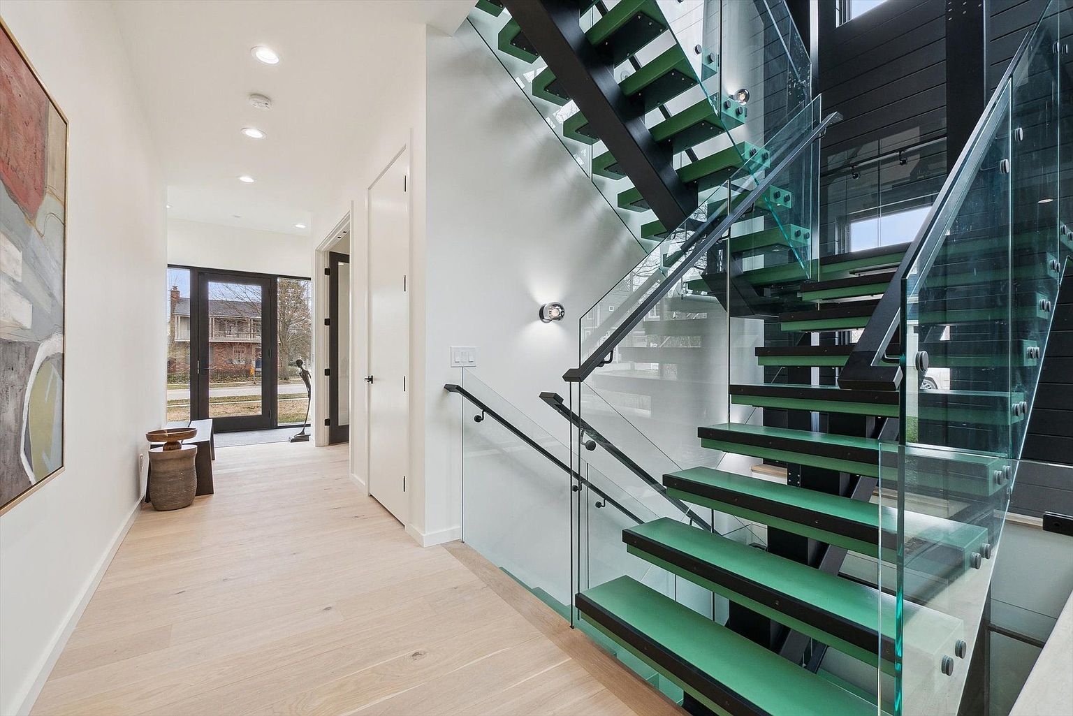 This interior shot showcases a modern hallway with a striking staircase. The staircase features green-tinted treads, black supports, and glass railings, creating a contemporary and open feel. The hallway has light wood flooring, white walls, and recessed lighting, leading to a doorway with a view to the outside.