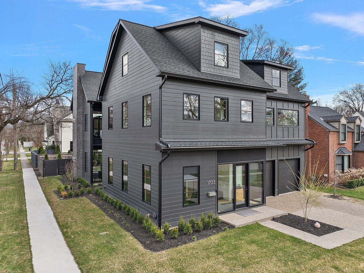 This is a front exterior view of a modern, multi-story home with a dark gray facade and black window frames. The house features a combination of horizontal siding and vertical accents, with a well-manicured lawn and landscaping. A paved driveway leads to an attached garage, and the overall impression is one of contemporary elegance and curb appeal.