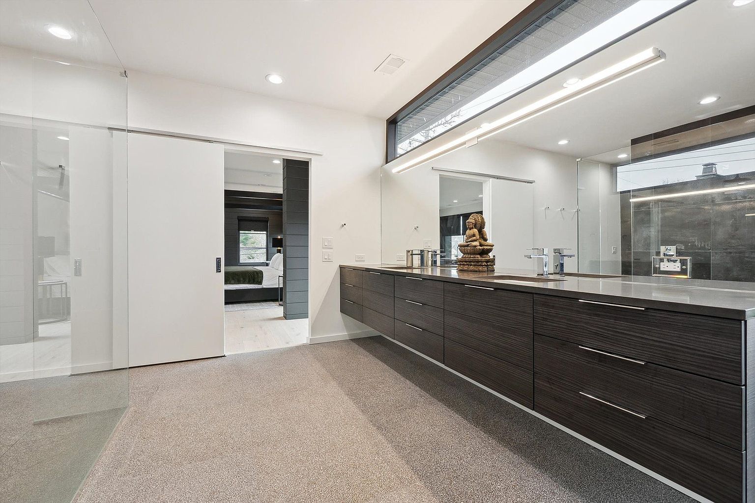 This is a modern primary bathroom featuring a long, floating vanity with dark wood-grain drawers and a light gray countertop. A large mirror stretches across the wall above the vanity, reflecting the light from a linear fixture above. The bathroom has a clean and minimalist aesthetic, with a glimpse into the bedroom through an open doorway.