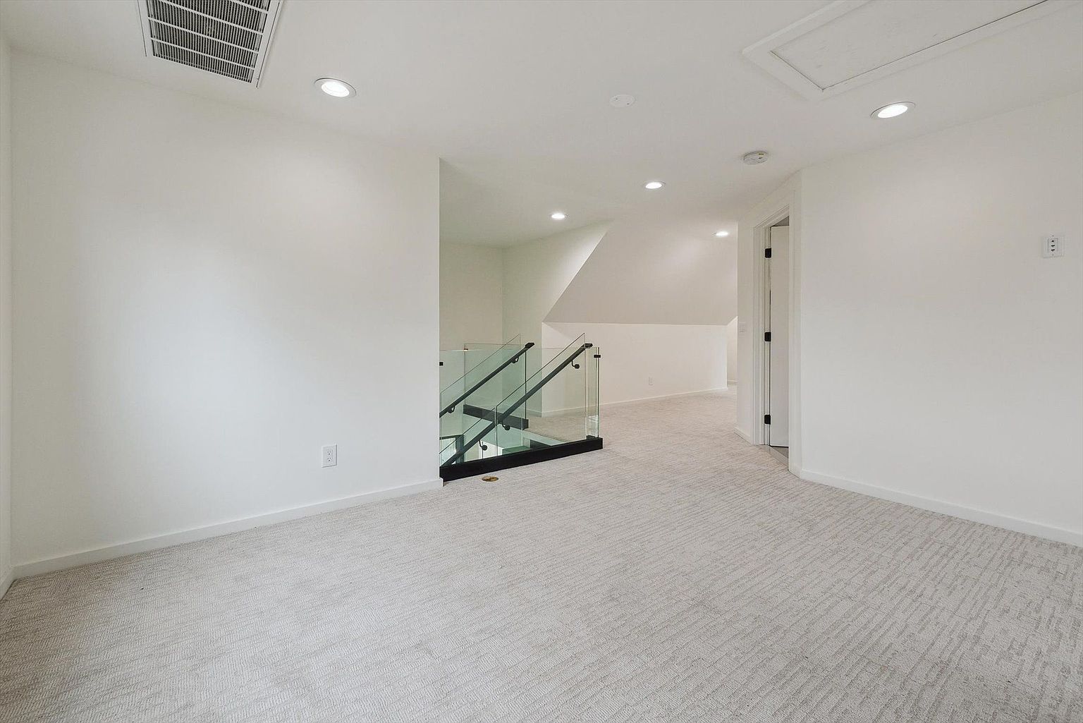 This is an interior shot of a hallway and stairs area, featuring a modern design with white walls and light gray carpet. A glass railing provides a sleek, open feel to the staircase. Recessed lighting illuminates the space, creating a bright and airy atmosphere.