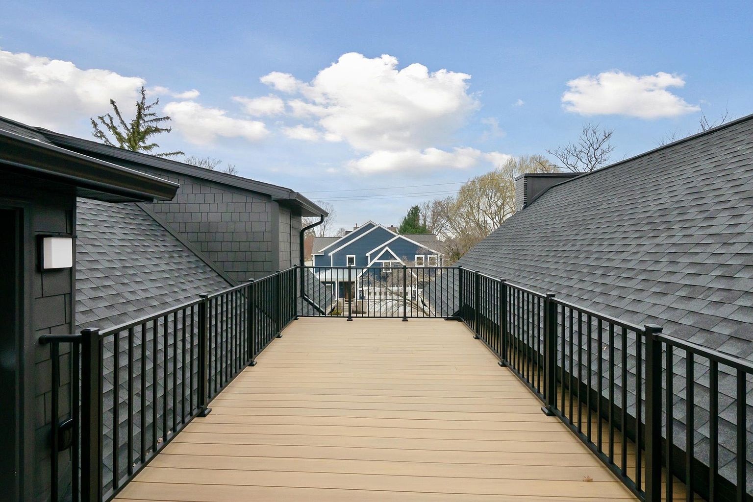 This image showcases a modern rooftop deck with black metal railings and a light brown composite deck surface. The deck is positioned between two houses with dark gray shingle roofing and siding, offering a unique vantage point. In the background, a blue house is visible, adding a pop of color to the scene under a partly cloudy sky.