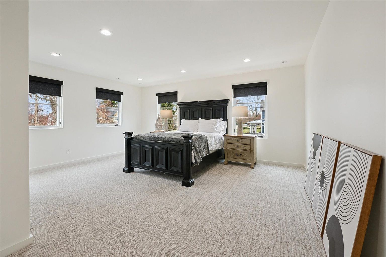 This is a spacious primary bedroom featuring a dark wood bed frame with white linens and a gray throw. The room is well-lit with natural light from multiple windows, which are adorned with dark blinds. The walls are painted white, and the floor is covered with a neutral-toned carpet, creating a bright and airy atmosphere.