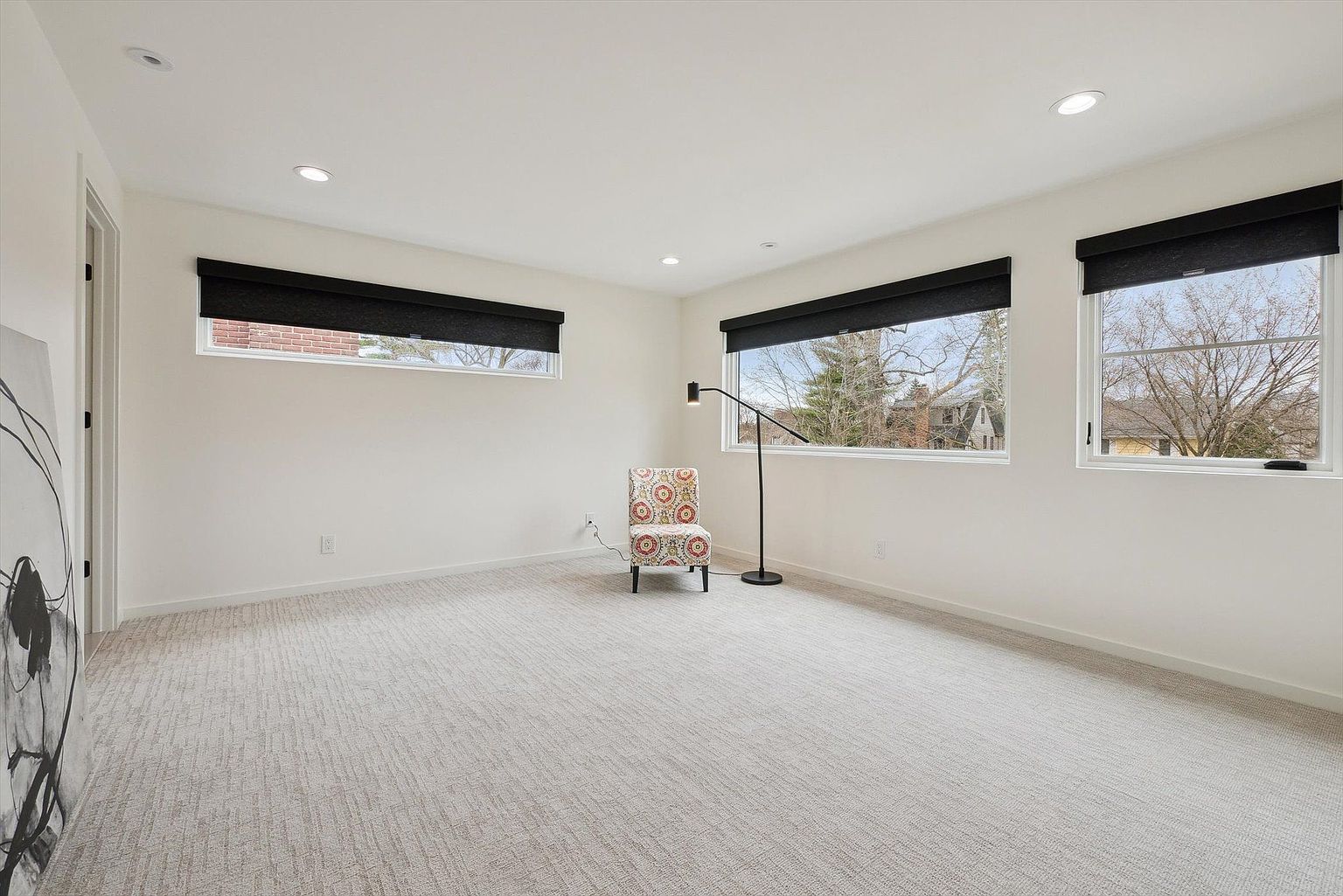 This is a bright and spacious bedroom featuring neutral-toned carpet and white walls, creating a clean and modern aesthetic. Three windows with black roller shades provide natural light and views of the outdoors. A patterned accent chair and a sleek black floor lamp add a touch of style to the minimalist design.