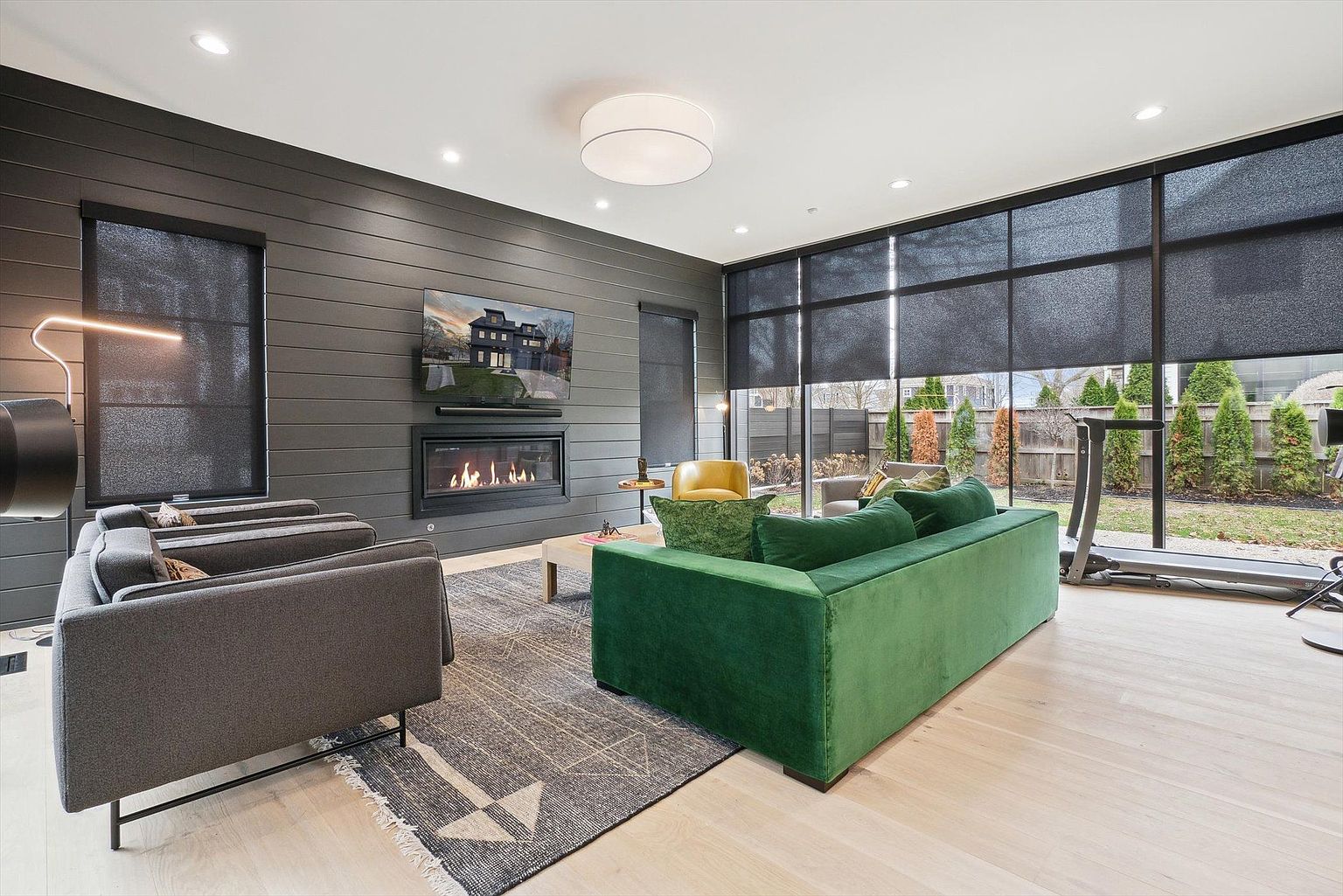This is a modern living room featuring a dark gray accent wall with a fireplace and a television mounted above it. The room is furnished with a green velvet sofa, gray armchairs, and a patterned area rug. Large windows with dark shades offer a view of the backyard, where a treadmill is visible, creating a seamless indoor-outdoor living experience.