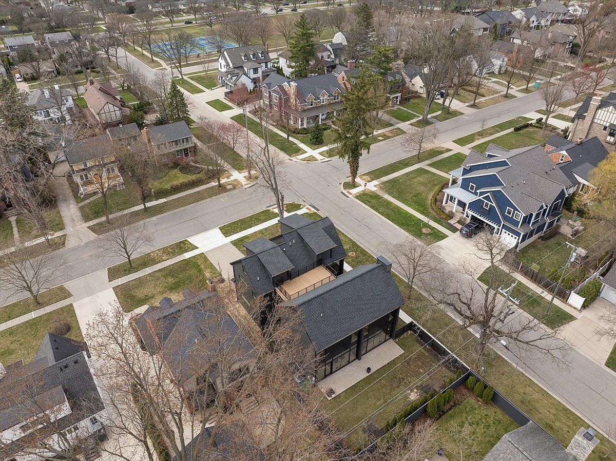 This aerial shot showcases a residential neighborhood with well-maintained homes and mature trees. The houses vary in architectural style, with some featuring dark roofs and modern designs, while others have a more traditional aesthetic. The manicured lawns and tree-lined streets contribute to the neighborhood's curb appeal.