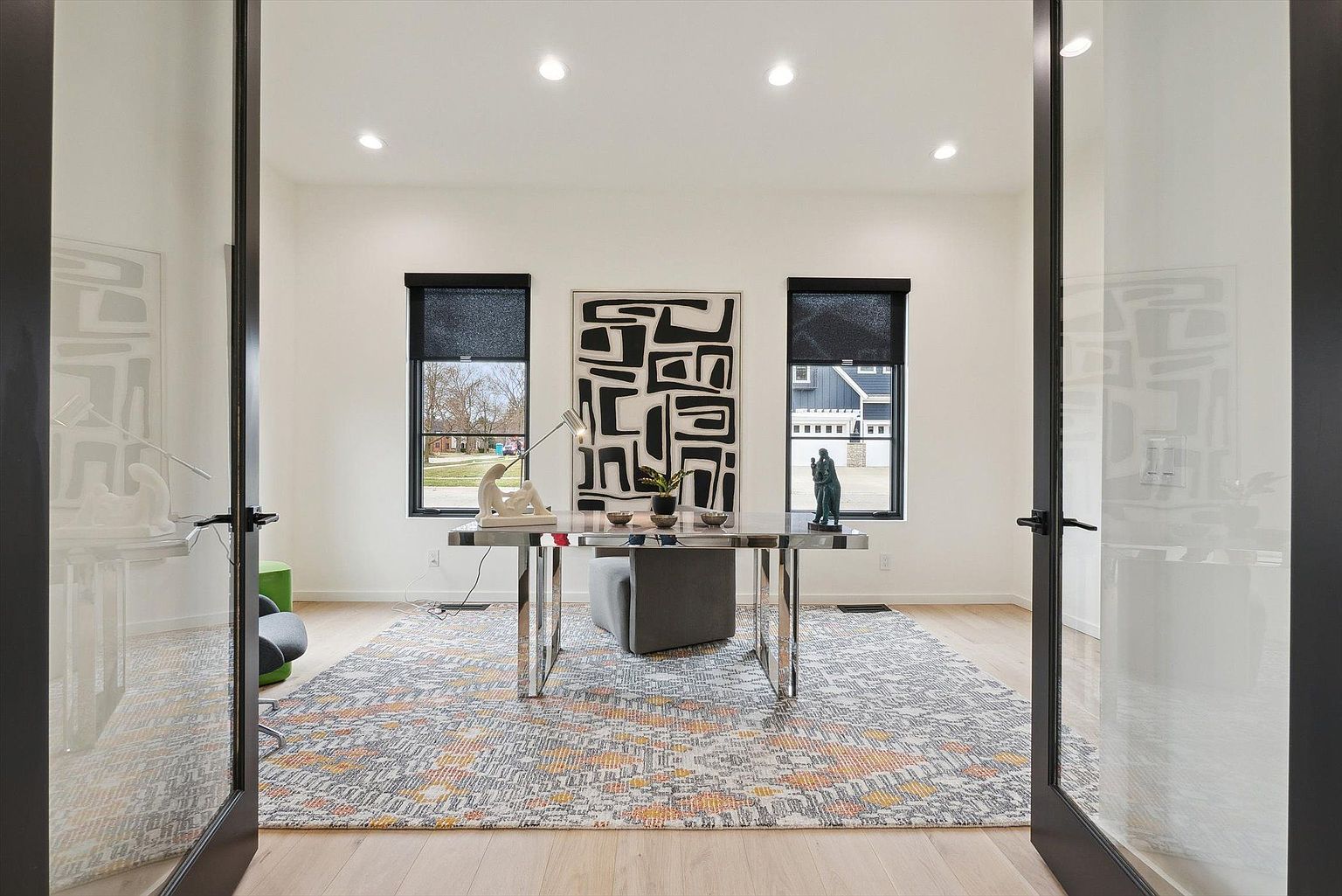 This is an interior shot of a modern office space. The room features a sleek desk with chrome legs, a gray ottoman, and a black and white abstract art piece. Natural light streams in through two windows with black shades, and the room is framed by open glass doors, creating an inviting and professional atmosphere.