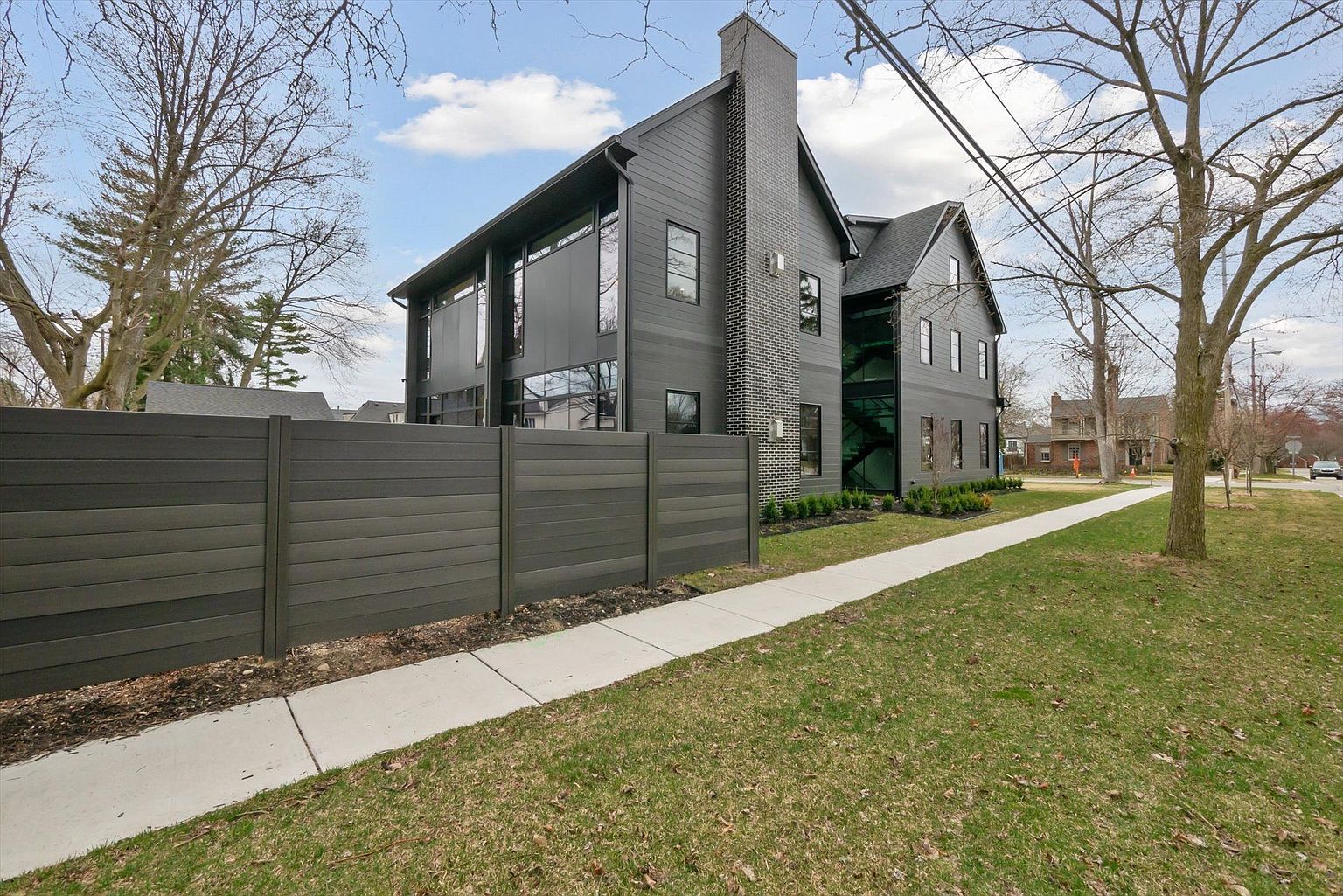 This is a side view of a modern, dark-colored house with a tall chimney and large windows. A dark horizontal fence runs along the property line, and a concrete sidewalk leads along the green lawn. The house features a unique architectural design with a combination of siding and brick elements.